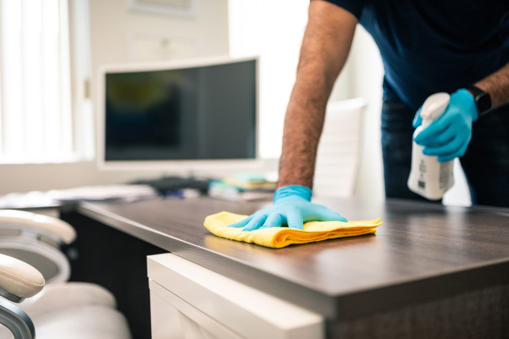 Cleaner disinfecting an office desk by wiping it down with a rag and spray