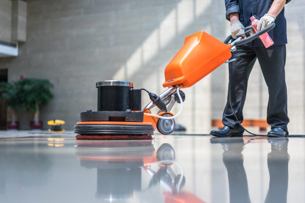 Working operating a floor care machine that is orange and black across a sleek floor with the machine reflecting off the floor